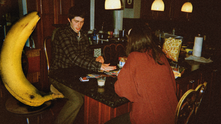 A photo a couple in a 1980s kitchen setting playing a card game with a really large banana while eating banana chips.