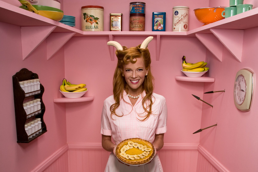 A stereotypical housewife with bananas in her hair, holds up a banana pie.
