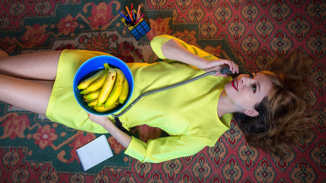 A lady in a yellow dress holds a bowl of bananas and talks on the phone.