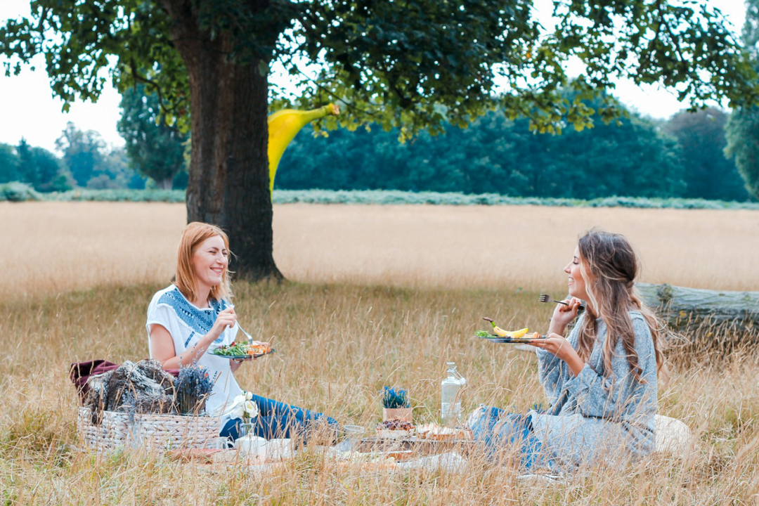 Two ladies enjoy a picnic as a banana watches from behind a tree.