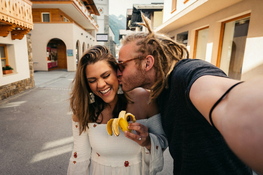 A man kisses his date while she tries to eat a banana.