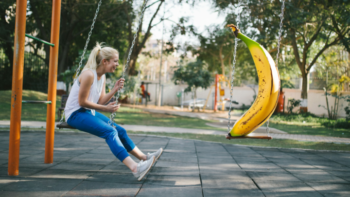 A girl laughs with a banana friend on swings in a playground.